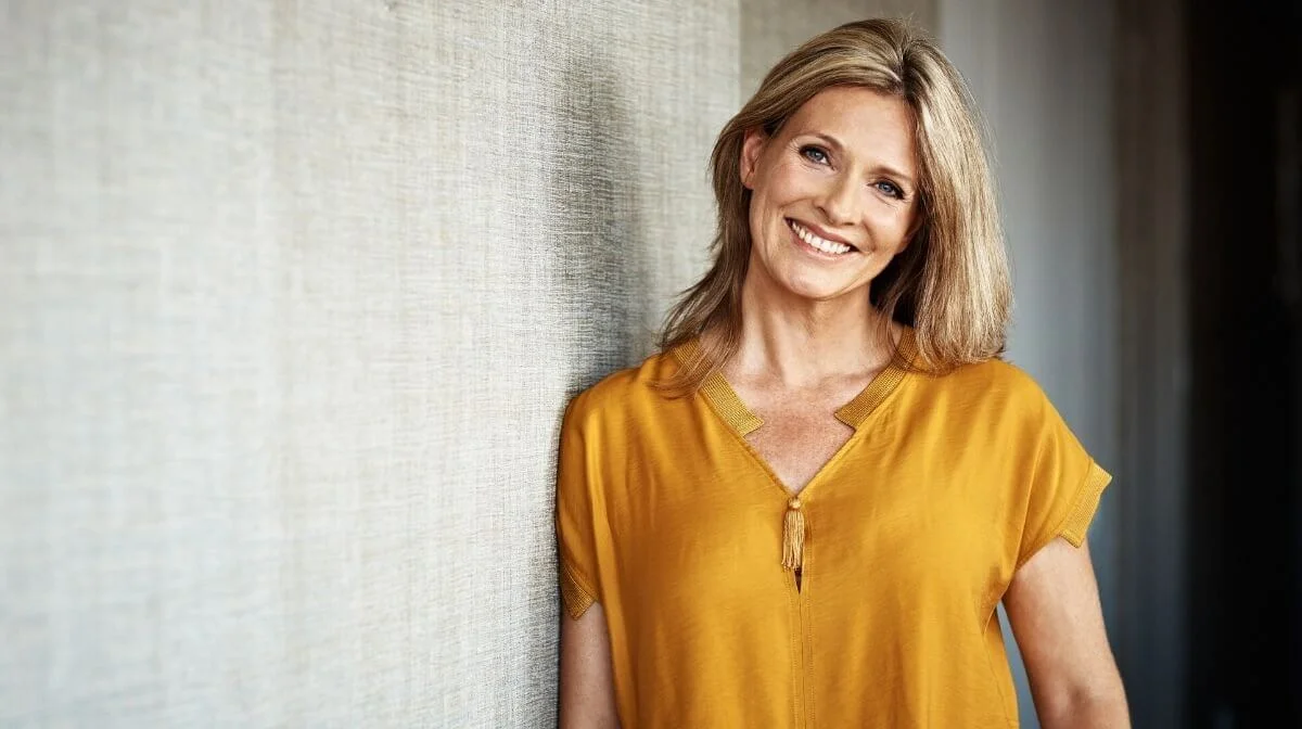 Woman with a yellow shirt leaning against a wall and smiling at camera